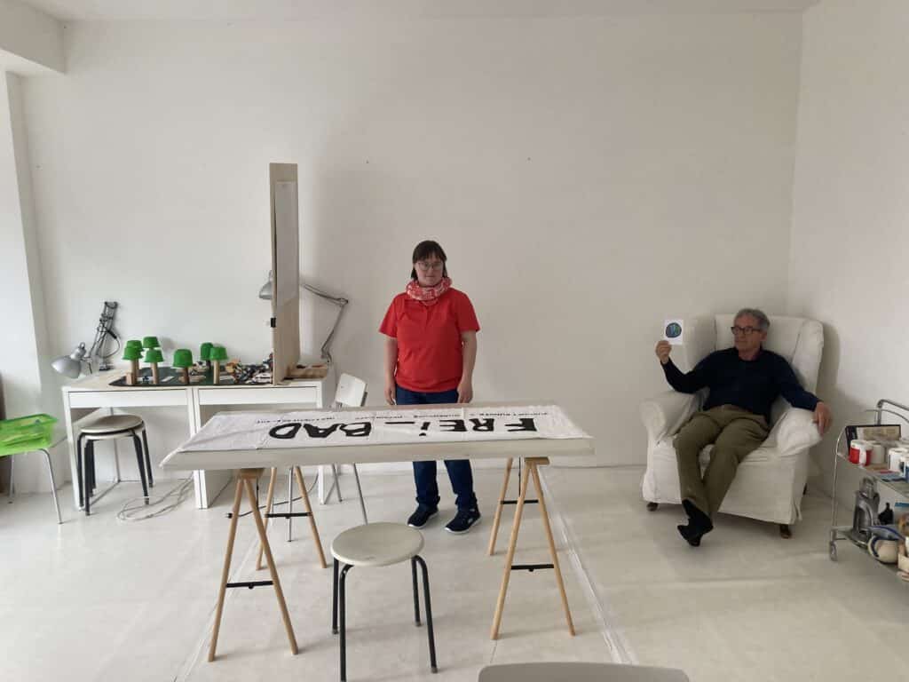 Ein weißer Raum. In der Mitte steht eine Frau mit rotem Oberteil und schaut in die Kamera. Rechts sitzt ein Mann in einem weißen Ohrensessel.