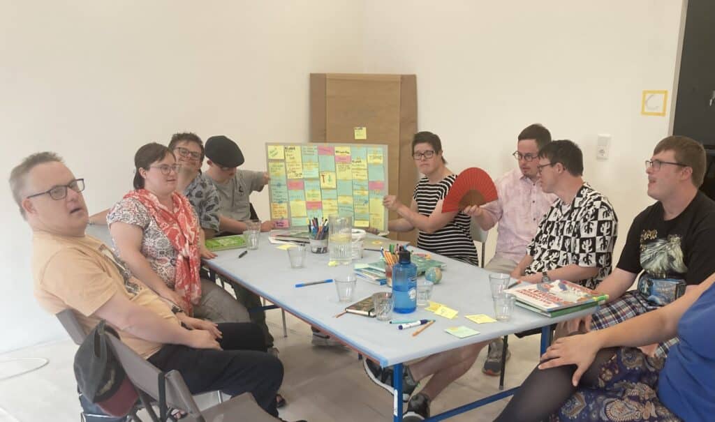 8 Menschen mit Down-Syndrom sitzen um einen Tisch. 2 halten eine Tafel mit farbigen beschrifteten Zetteln hoch..