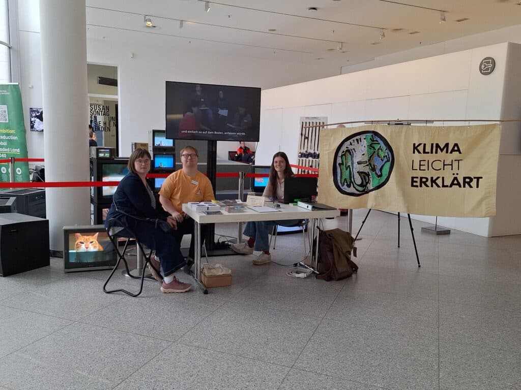 Ein Tisch in einem hohen Raum. Es ist das Foyer der Bundeskunsthalle in Bonn. An einem Tisch sitzen 2 Frauenn und1Mann.. Hinter ihnen ist ein Bildschirm und das Banner von Klima Leicht Erklärt.
