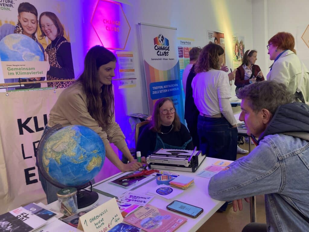 Eine trübselige Stimmung, viele Menschen sind zu sehen. Vorne ist ein Tisch. 2 Frauen stehen dahinter, ein Mann kniet davor. Sie sind im Gespräch.