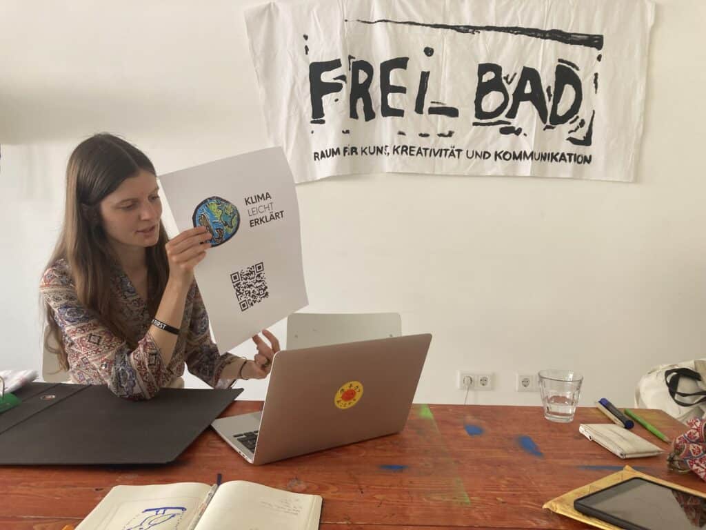 Eie Frau zeigt ein A3 großes Blatt Papier. Vor ihr ist ein aufgeklappter Laptop auf dem Tisch.