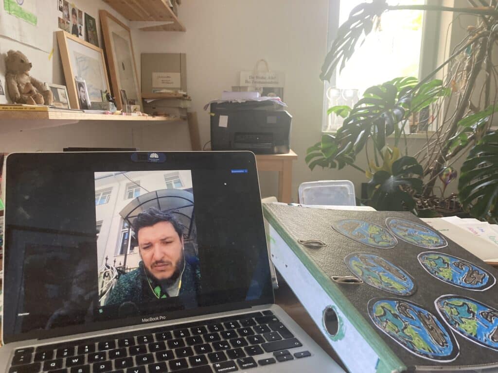 Ein aufgeklappter Laptop, ein Mann ist auf dem Bildschirm zu sehen. Neben dem Laptop liegt ein Aktenordner. Er ist mit mehreren Weltkugeln beklebt. Im Hintergrund ist ein Kopierer und eine Monstera Pflanze zu sehe.