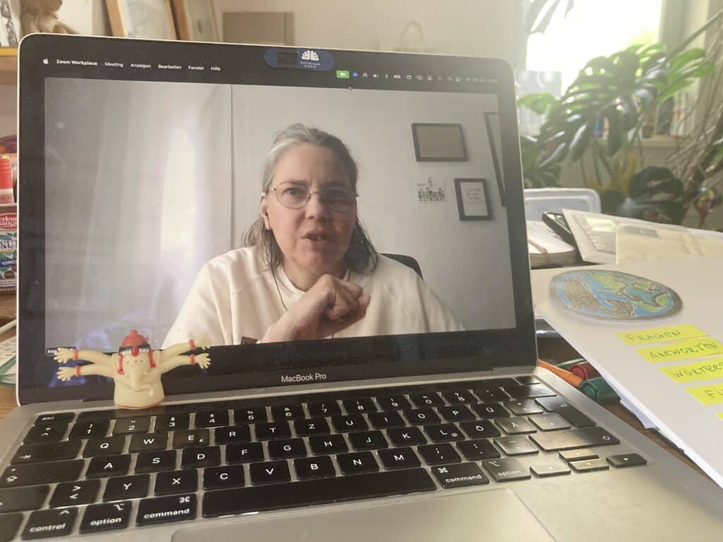 Ein aufgeklappter Laptop, eine Frau mit weißem Sweatshirt st auf dem Bildschirm zu sehen. Neben dem Laptop liegt Büromaterial.. Im Hintergrund ist eine Monstera Pflanze zu sehen.