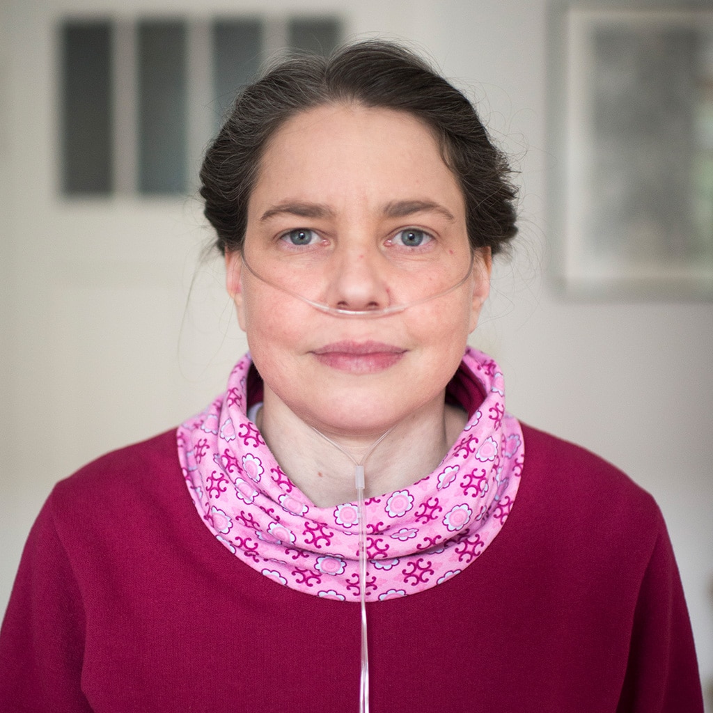 Eine junge Frau blickt in die Kamera. Sie trägt ein rotes Oberteil mit rosa Kragen. Durchsichtige Schläuche sind sichtbar. Der Schauch führt unter der Nase entlang, dann hinter beide Ohren. Unter ihrem Kinn kommen beide Schläuche wieder zusammen und gehen nach unten. Das bedeutet: Sie atmet zusätzlichen Sauerstoff ein.