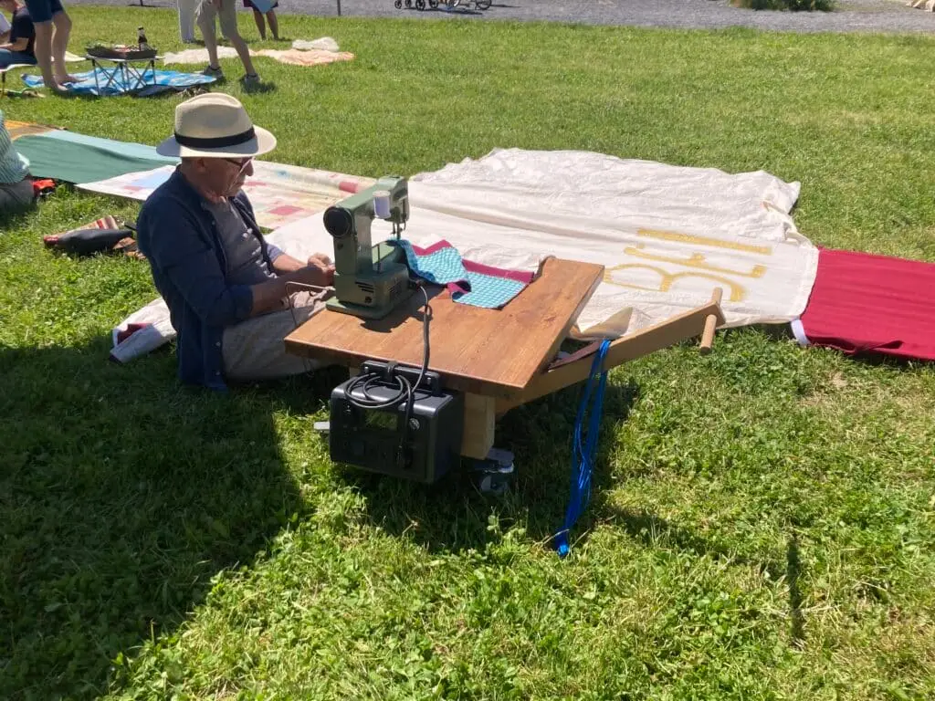 Ein Mann mit Strohhut sitzt vor einer Nähmaschine. Er näht ein Stück Stoff an eine lange bunte Picknick Decke, die auf einer Wiese liegt.