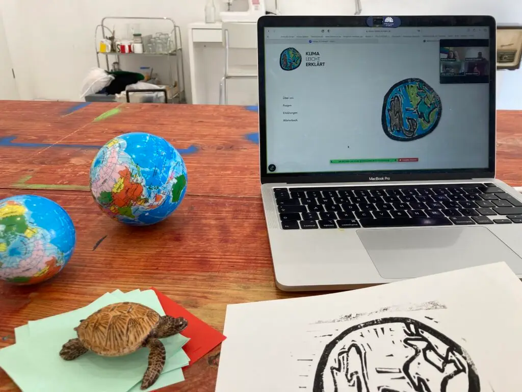 Ein Holztisch, darauf steht ein offener Laptop, auf dem Bildschirm sieht man die Klima Leicht Erklärt Internetseite. Auf dem Tischliegen 2 Bälle, bedruckt wie eine Weltkugel. Davor ist eine Wasserschildkröte aus Plastik.