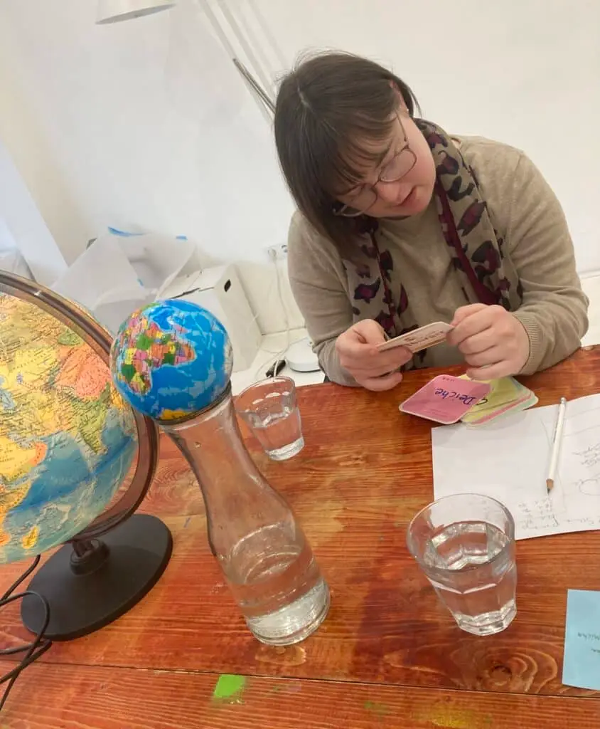 Eine Frau schaut sich einen einen Bierdeckel an, vor ihr liegen noch 2 auf dem Tisch. 2 Gläser mit Wasser und eine Karaffe stehen auf dem Tisch, daneben 2 Globen.