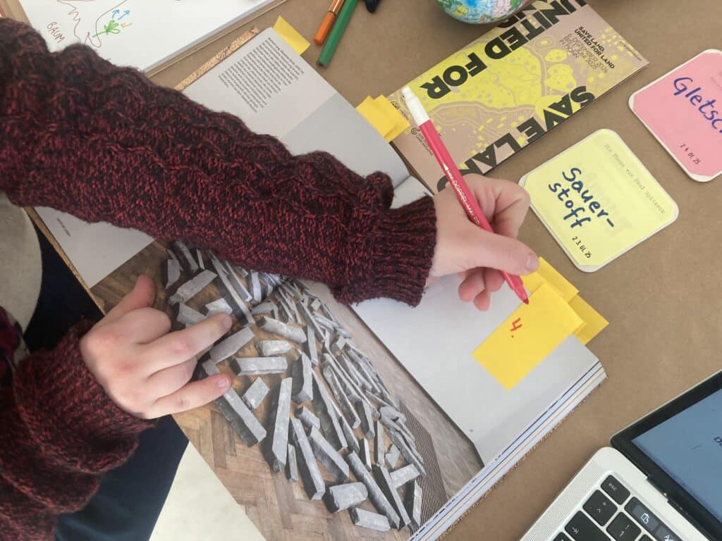 Perspektive von oben auf einen Schreibtisch. Eine linke Hand schreibt die Zahl 4 auf einen gelben Post-it, geklappt auf einer Katalogseite. Zu sehen ist ein Foto eines Steinkreises aus Basalt.