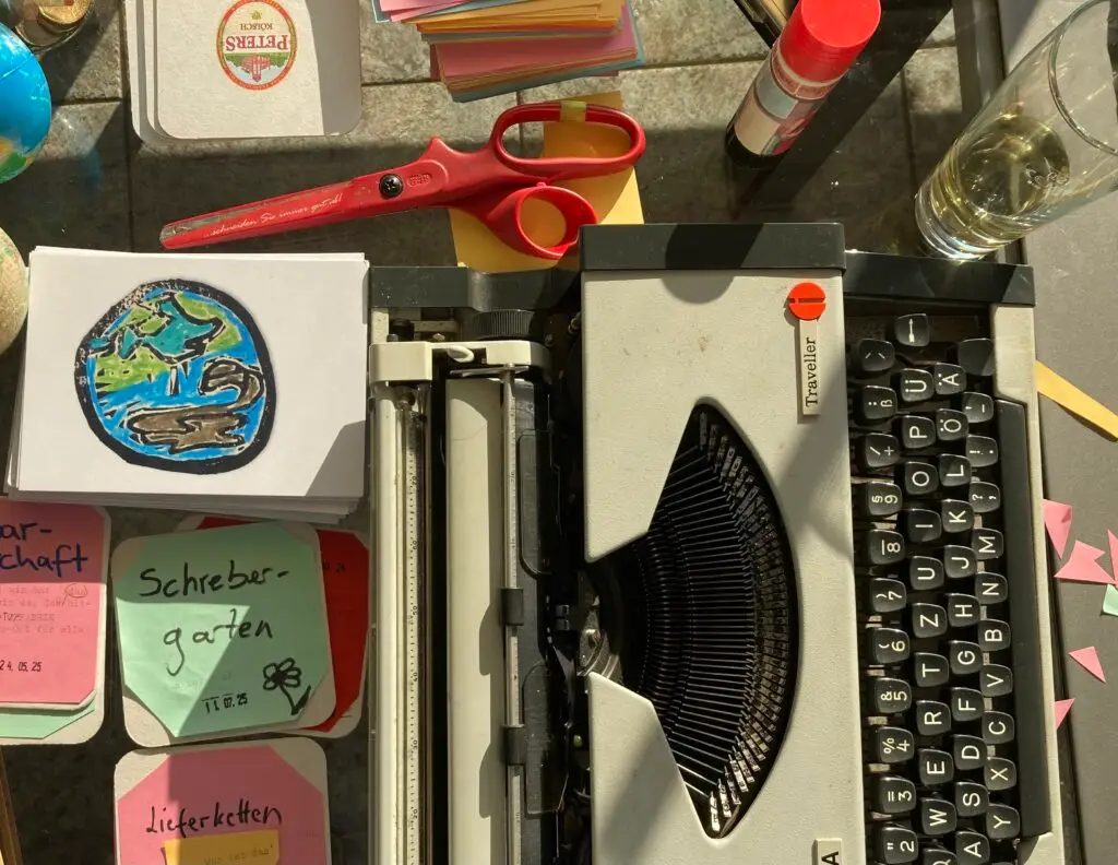 Blick von oben: Eine mechanische Schreibmaschine und 2 Bierdeckel mit den Wörtern "Schrebergarten" und "Lieferketten". Daneben liegen eine Schere und Karten mit einer Weltkugel.