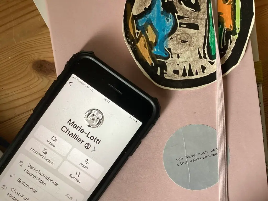 Ein Handy mit dem Kontakt von Marie-Lotti, ihr Profilbild ist ein Hund. Das Handy liegt auf einem rosa Buch. Darauf klebt ein kleiner grauer Sticker "ich habe den wind wahrgenommen". Daneben ein Linoldruck einer Weltkugel, mit Buntstiften angemalt: unser Logo.