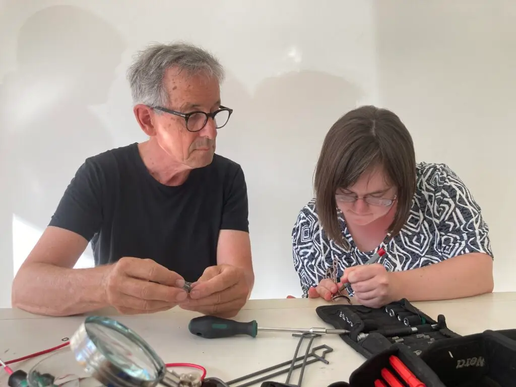 Ein Mann und eine Frau sitzen an einem Tisch. Die Frau benutzt einen Schraubendreher. Auf dem Tisch liegt Werkzeug.