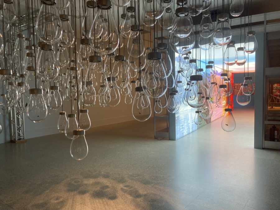 Ein Musezmsraum mit hellgrauem Boden. Von der Decke hängen unzählige Glühbirnen. Rechts hinten ist der Durchgang zum nächsten Raum.