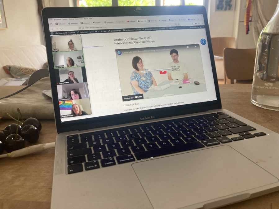 Ein aufgeklappter Laptop, rechts steht ein Wasserglas, links liegen 4 rote Kirschen. Auf dem Bildschirm ist eine Zoom Konferenz zu sehen.