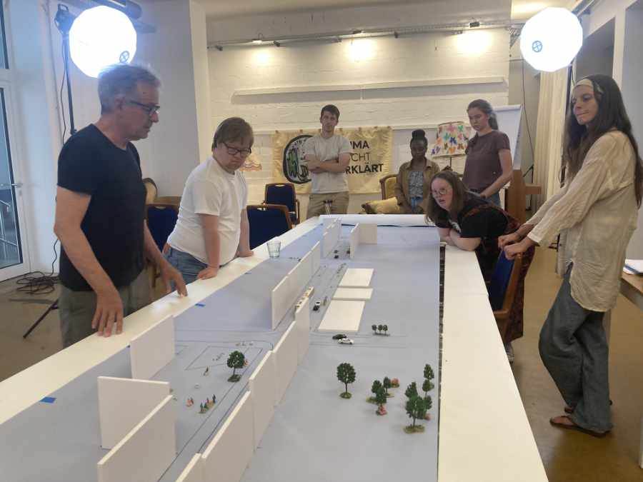 7 Menschen stehen um einen weißen langen Tisch. Sie schauen sich ein Modell einer Straße an, mit kleinen Autos, Bäumen, Fahrrädern und Menschen.