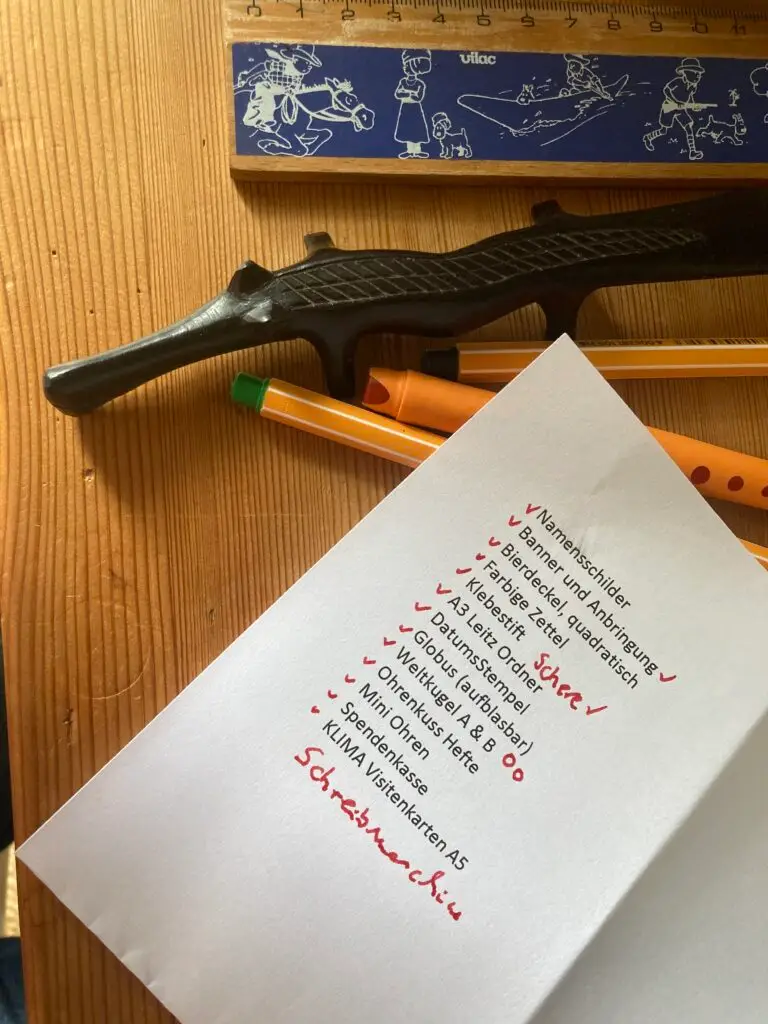 Ein Liste, mit der Schreibmaschine geschrieben. Die erledigten Dinge sind mit rotem Stift abgehakt. Der Zettel liegt auf einem Schreibtisch aus Holz, daneben ein Lineal mit Tim und Struppi Motiven, ein Brieföffner in Form eines Krokodils und 3 Stifte