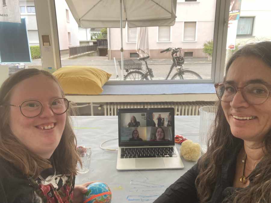 2 Frauen schauen die Kamera. Sie lächeln. Zwischen ihnen steht ein Laptop. Auf dem Bildschirm sind mehrere Menschen zu sehen - sie zoomen.