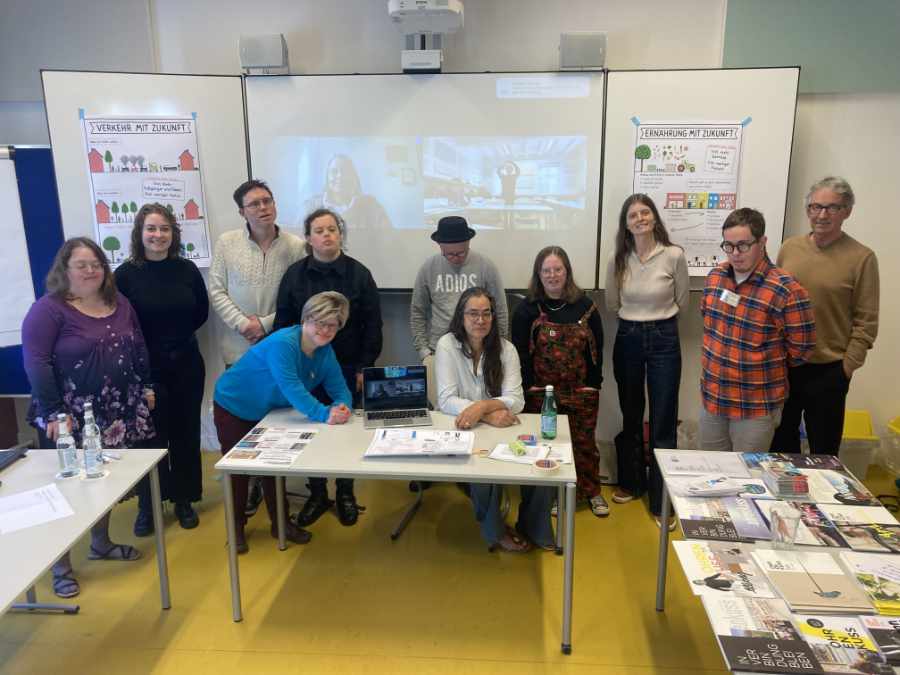 11 Menschen stehen und sitzen in einem Seminarraum vor der Tafel. Auf der Tafel ist etwas projiziert: Eine Frau. Links und rechts sind Plakate auf die Tafel geklebt.