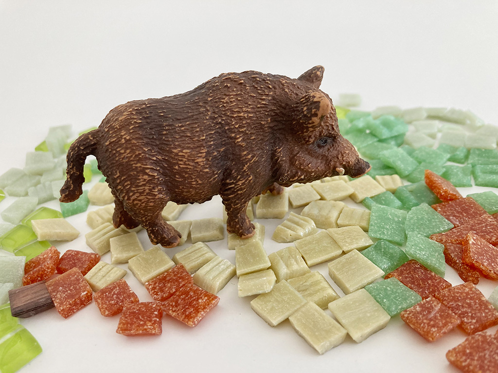 Auf dem Foto sieht man eine Miniatur von einem Wildschwein. Es steht auf Mosaik Steinen aus verschiedenen Farben.