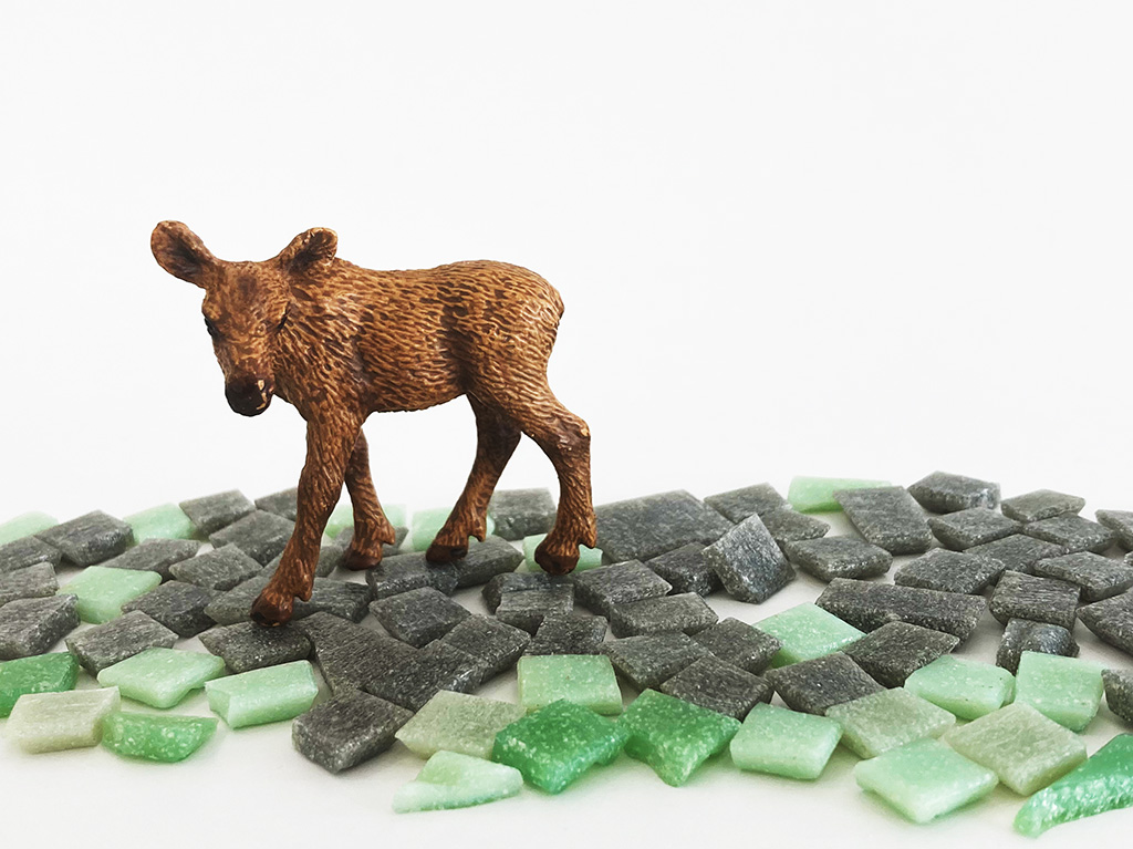 Auf dem Foto sieht man die Miniatur von einem Baby Elch. Er steht auf schwarzen und grünen Mosaik Steinen.