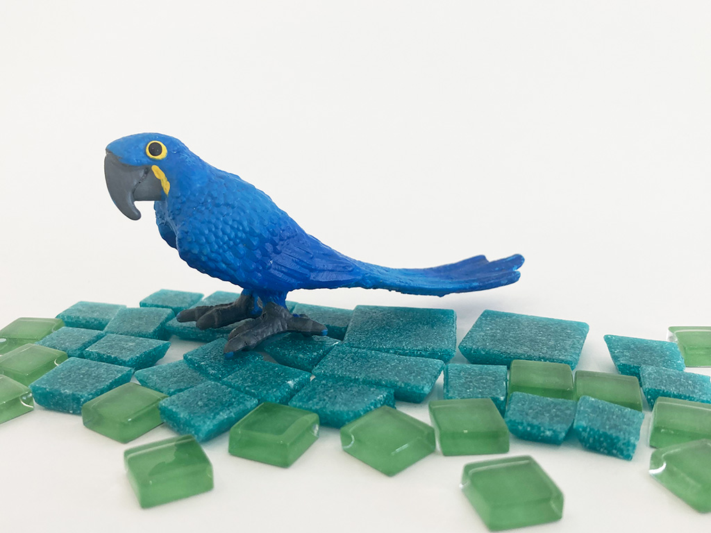 Auf dem Foto sieht man eine Miniatur von einem blauen Papageien. Er steht auf grünen und und blauen Mosaik Steinen.
