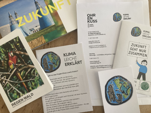 Es sind viele Informationsmaterialien zu sehen. Man kann gut das Logo von KLIMA leicht erklärt erkennen. Es gibt eine Karte mit einem Papagei und dem Schriftzug Regenwald. Und eine Ohrenkuss Ausgabe mit dem Schriftzug Zukunft.