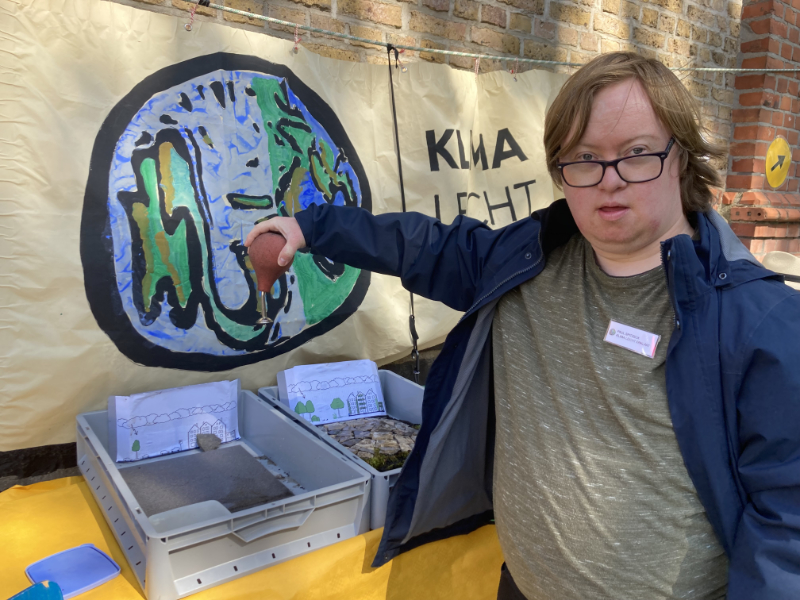 Ein junger Mann steht vor einem Info Tisch. Er zeigt auf das Banner. Auf dem Banner ist die Weltkugel zu sehen.