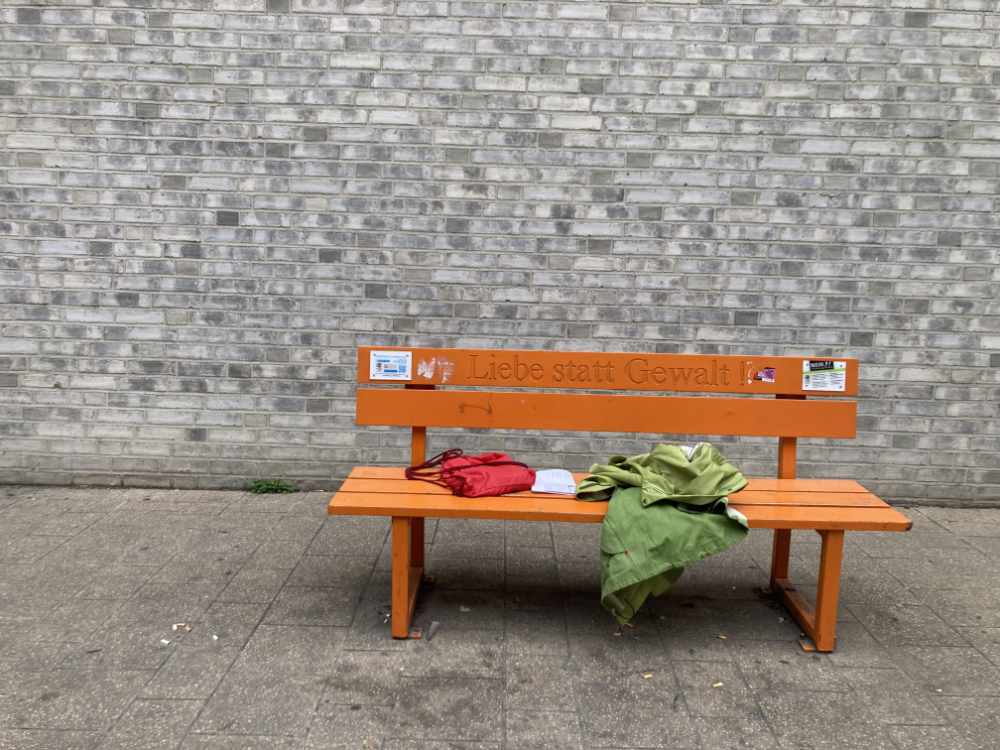 Eine orangene Bank, darauf steht: Liebe statt Gewalt. Auf der Bank liegen ein roter Rucksack, weißes Papier und eine grüne Jacke.