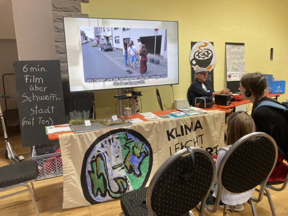 Ein Informationsstand ist zu sehen. Vorne kann man ein Banner sehen, mit dem Logo von KLIMA leicht erklärt. Hinter dem Tisch wird ein Film gezeigt, zum Thema Schwammstadt. Zwei Personen schauen sich den Film an. Sie tragen Kopfhörer.